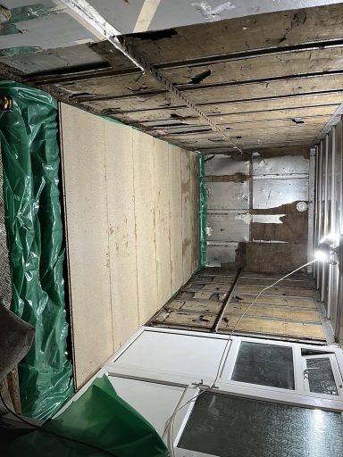 Empty room undergoing renovation, featuring bare walls and a plywood floor.